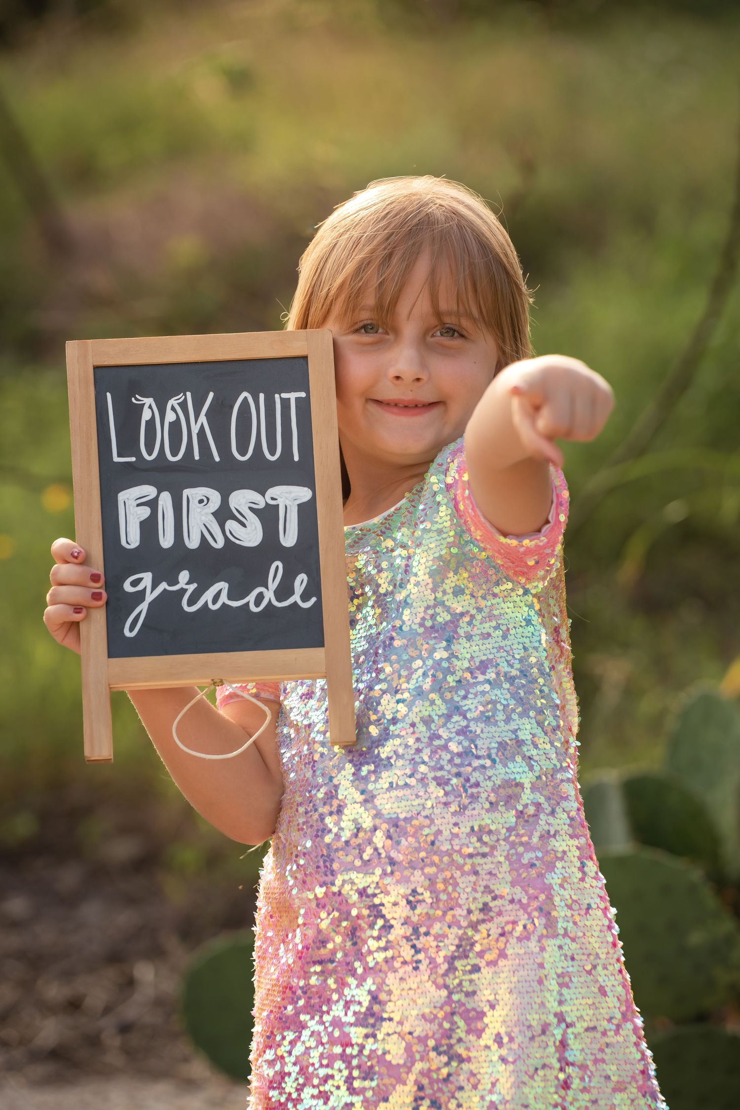 Young girl in sequin dress holds chalkboard sign reading Look Out First Grade while pointing at camera outdoors.
