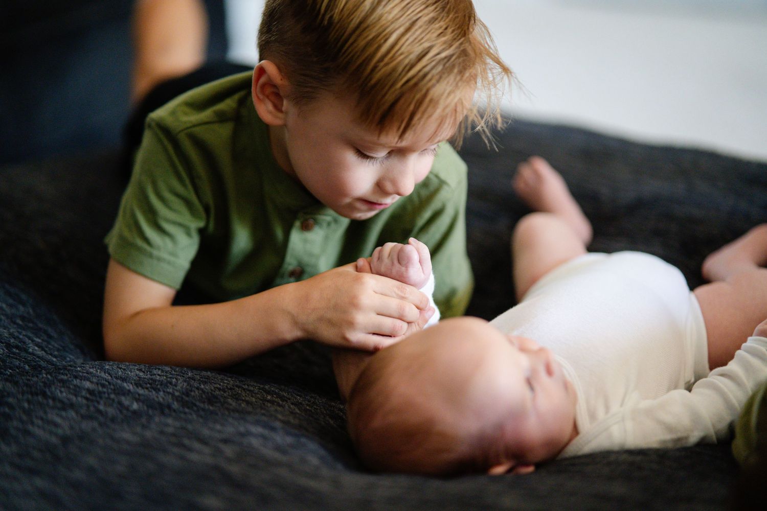 Young boy in green shirt gently holding newborn baby's hand while lying on a dark sofa.