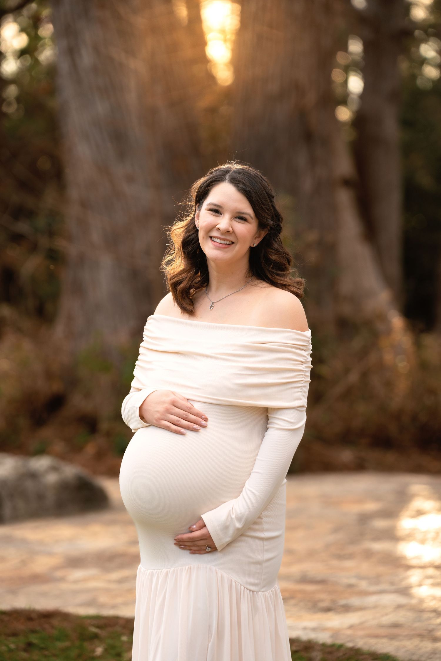 Smiling pregnant woman in white off-shoulder gown cradling her baby bump outdoors at golden hour.