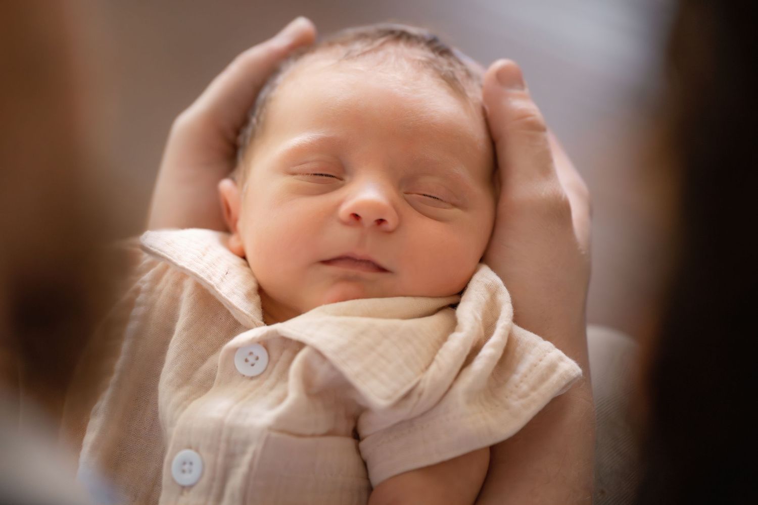 Sleeping newborn baby cradled gently in adult hands, wearing a beige onesie, with a peaceful expression.