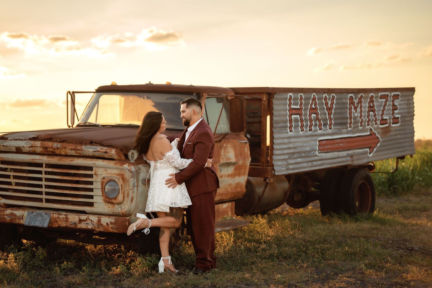 Couple poses romantically by a rustic vintage truck with Hay Maze sign at golden sunset in a field.