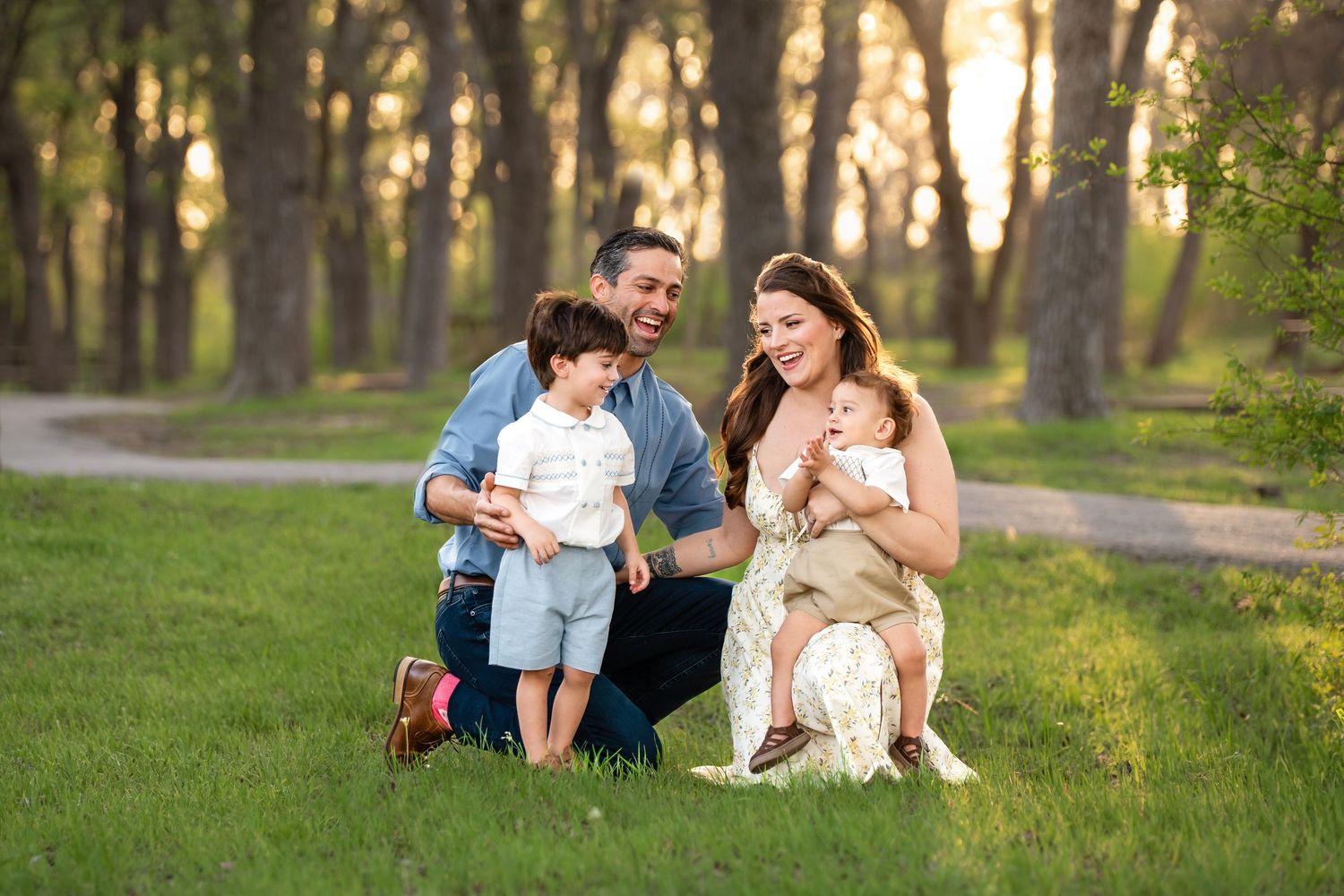 Family of four smiling outdoors in a sunlit park, parents with two young children on green grass among trees.