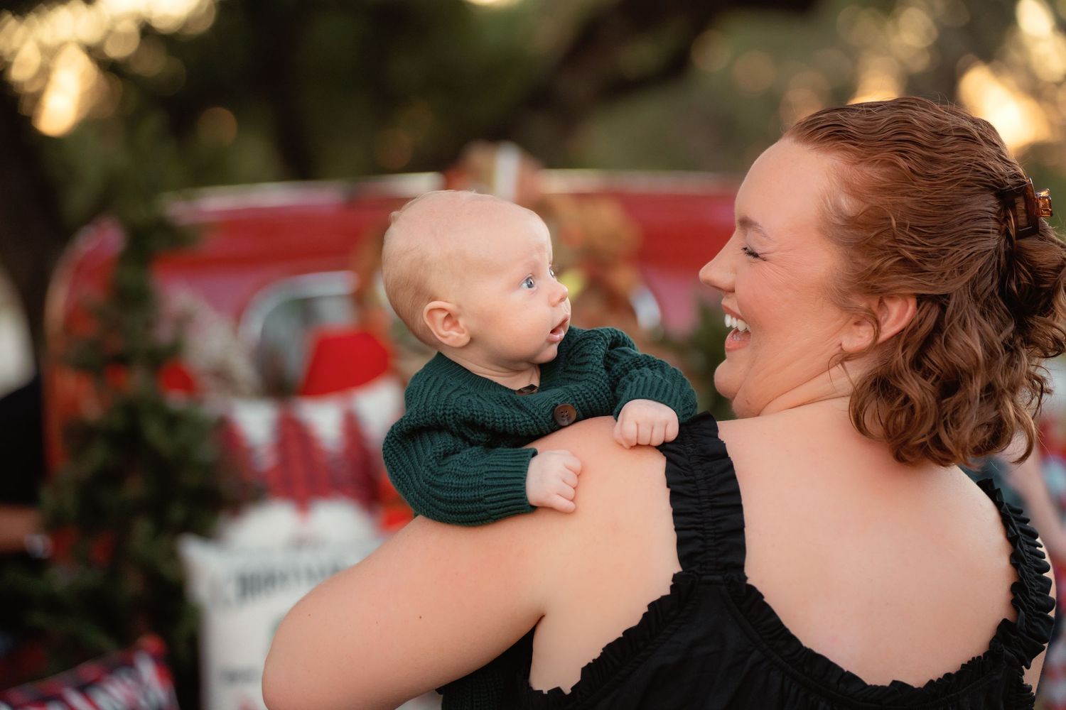 A smiling young woman with red hair holds a baby in a green outfit outdoors near a red truck during a holiday photo session.