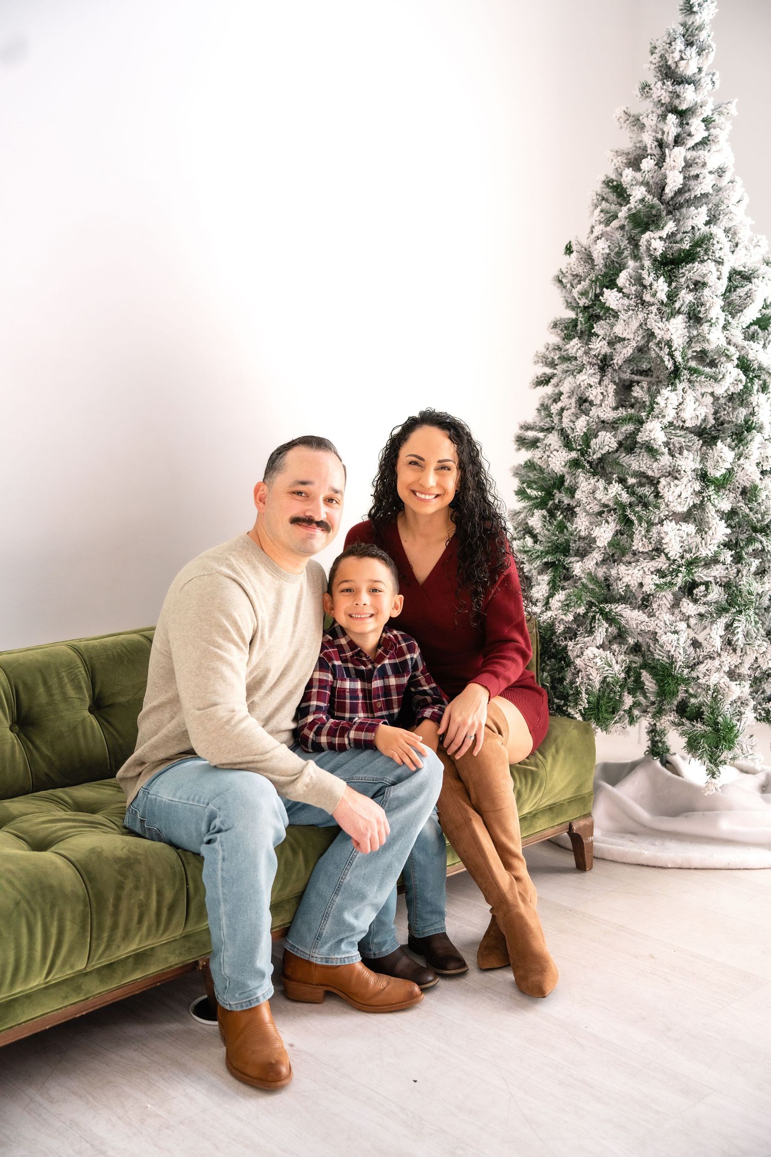 Family sitting and laughing together on green couch with festive Christmas tree in background during holiday photo shoot.