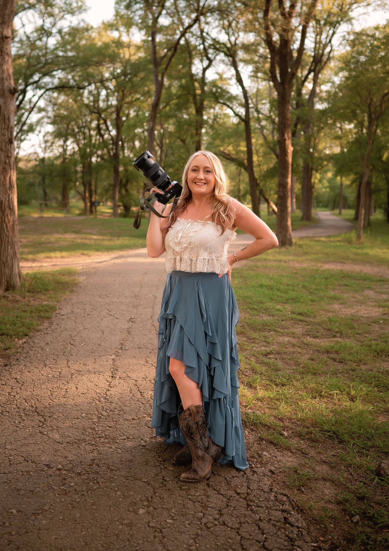 Smiling blonde woman in lace top, blue ruffle skirt, and cowboy boots holding a camera on a dirt path.