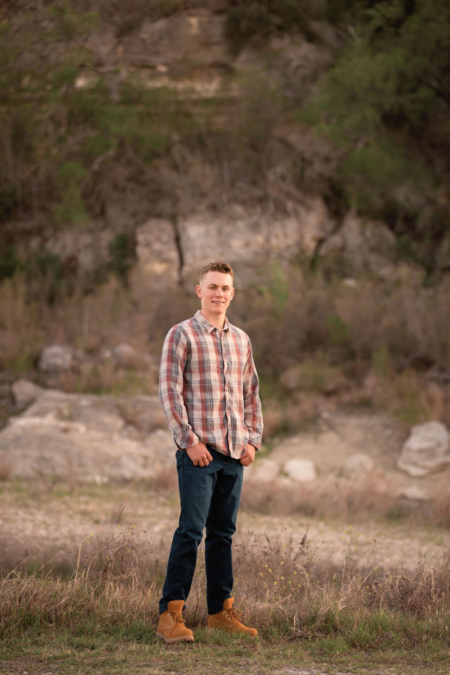Young man in plaid shirt and jeans posing outdoors near rocky terrain and green trees in natural light.