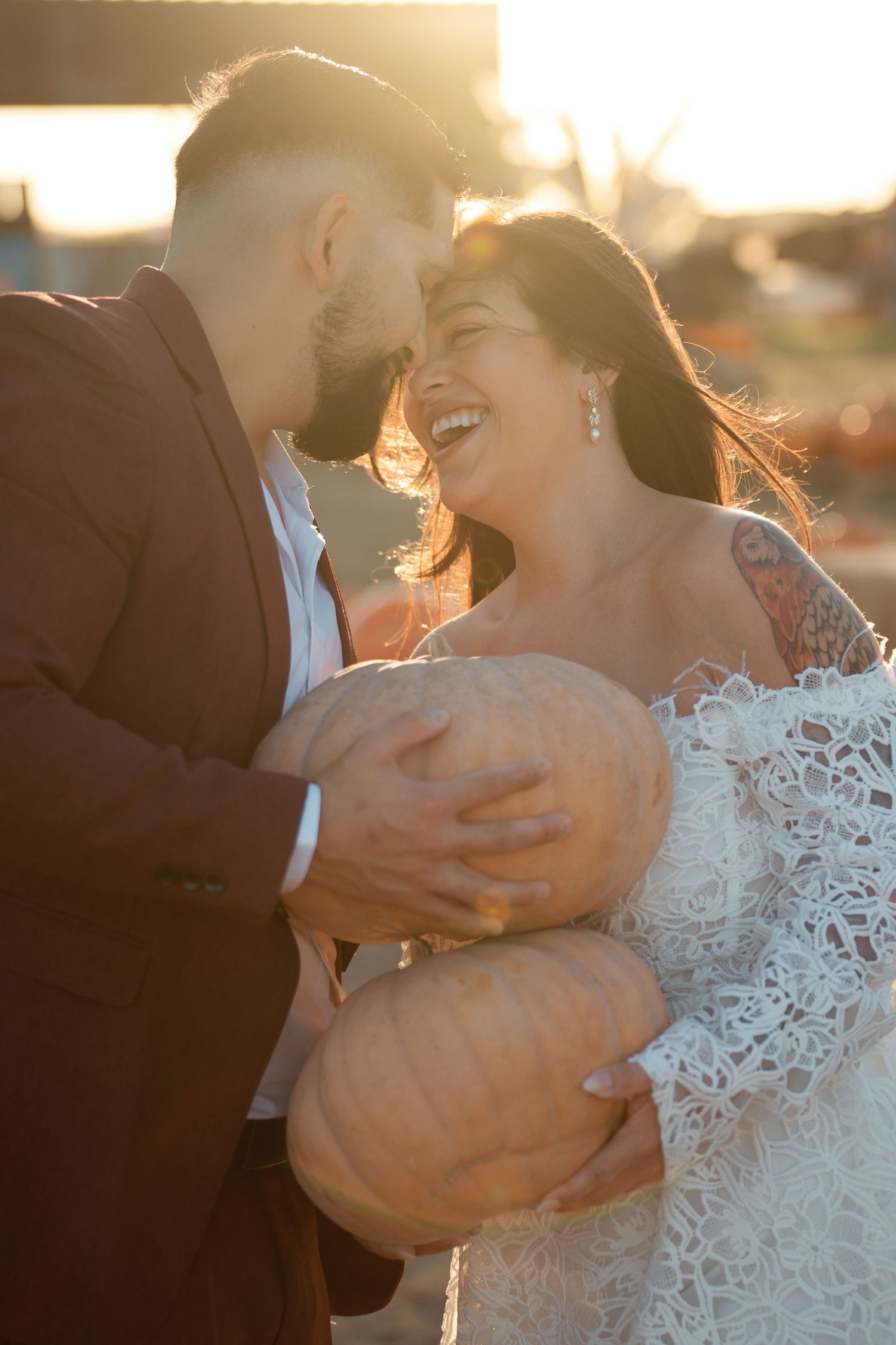 A joyful bride and groom share a laugh while holding pumpkins together in golden sunset light.