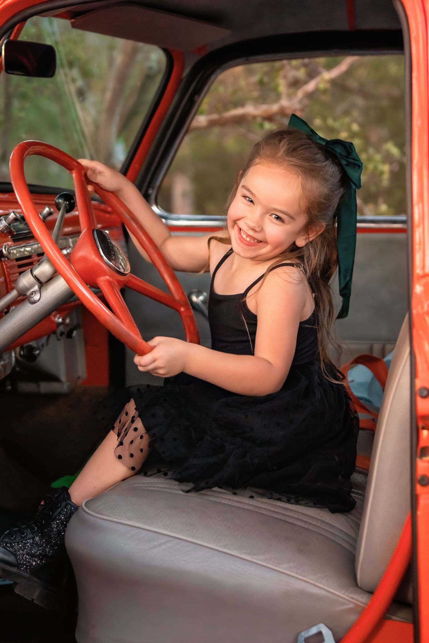 Smiling young girl in black dress sitting behind red steering wheel of vintage orange truck.