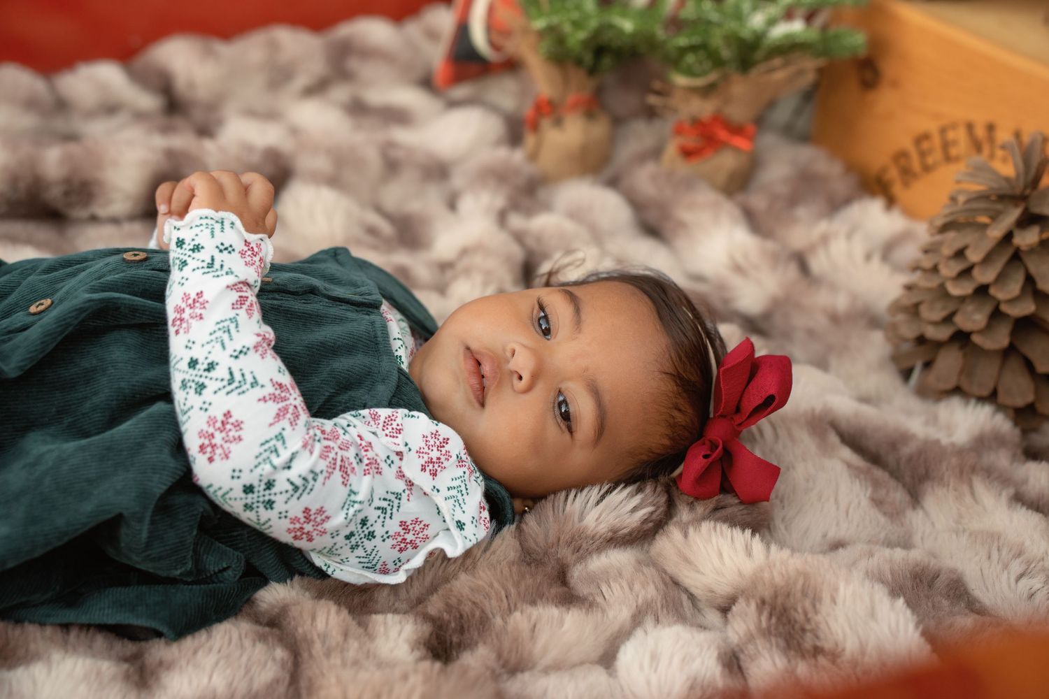 Baby girl in green dress with floral sleeves and red bow lies on fur blanket in festive Christmas setting.