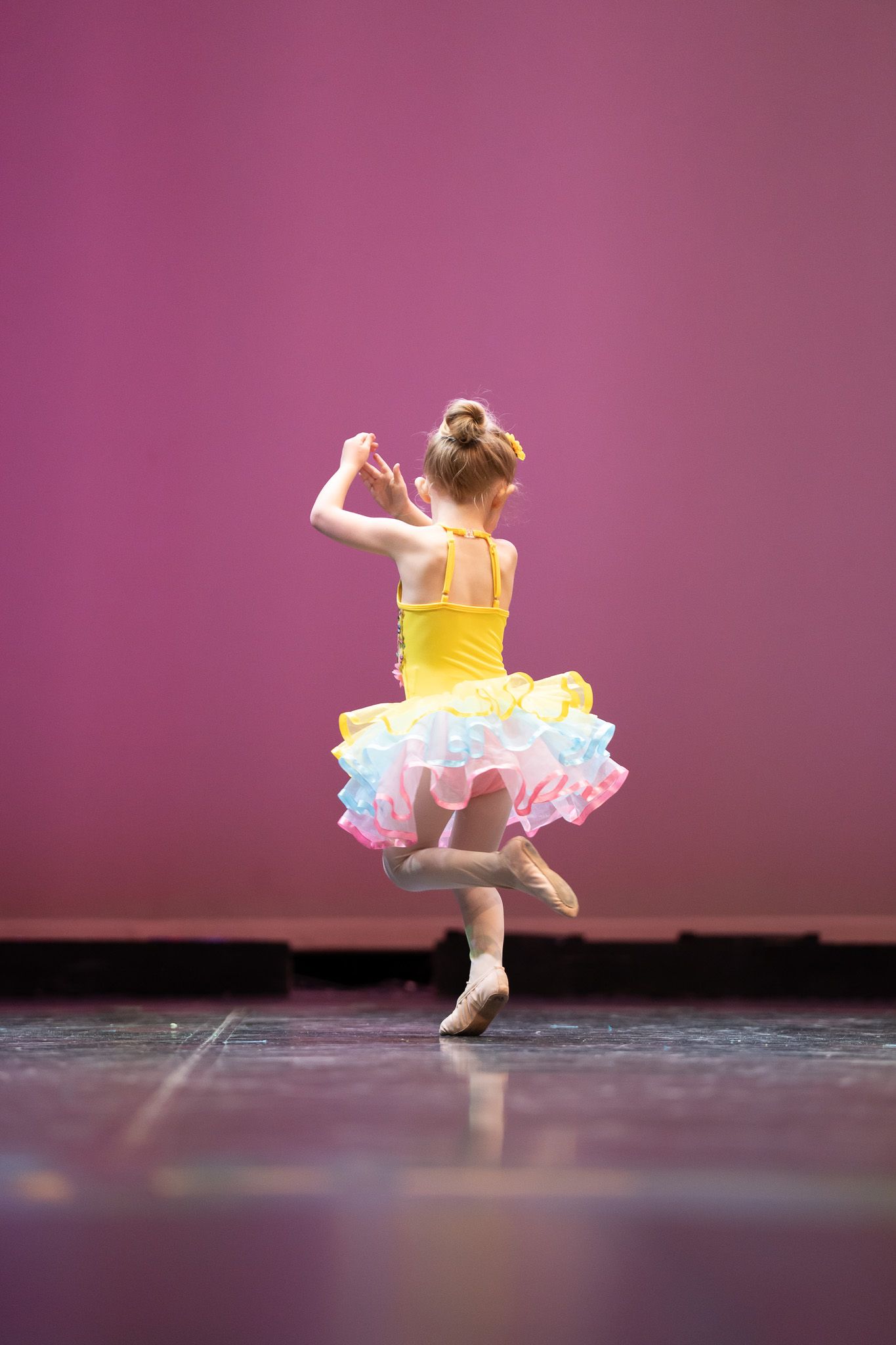 Young dancer in yellow tutu performing graceful movements against a purple backdrop.