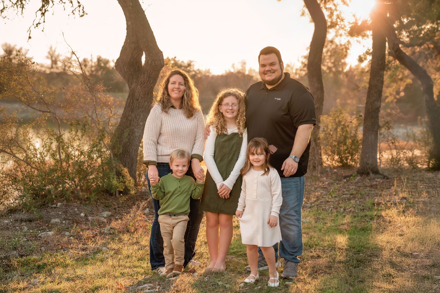 Family of five posing outdoors at golden hour among trees with warm sunlight glowing in the background.