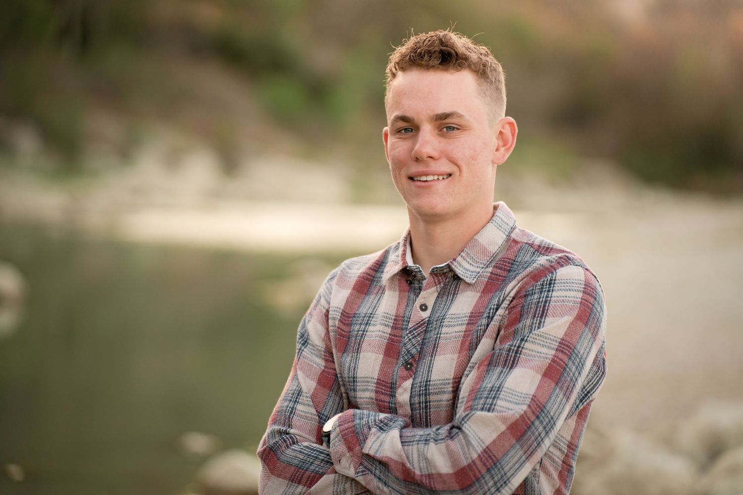 Young man with crossed arms smiling outdoors near a river, wearing a plaid shirt in a senior portrait photo.