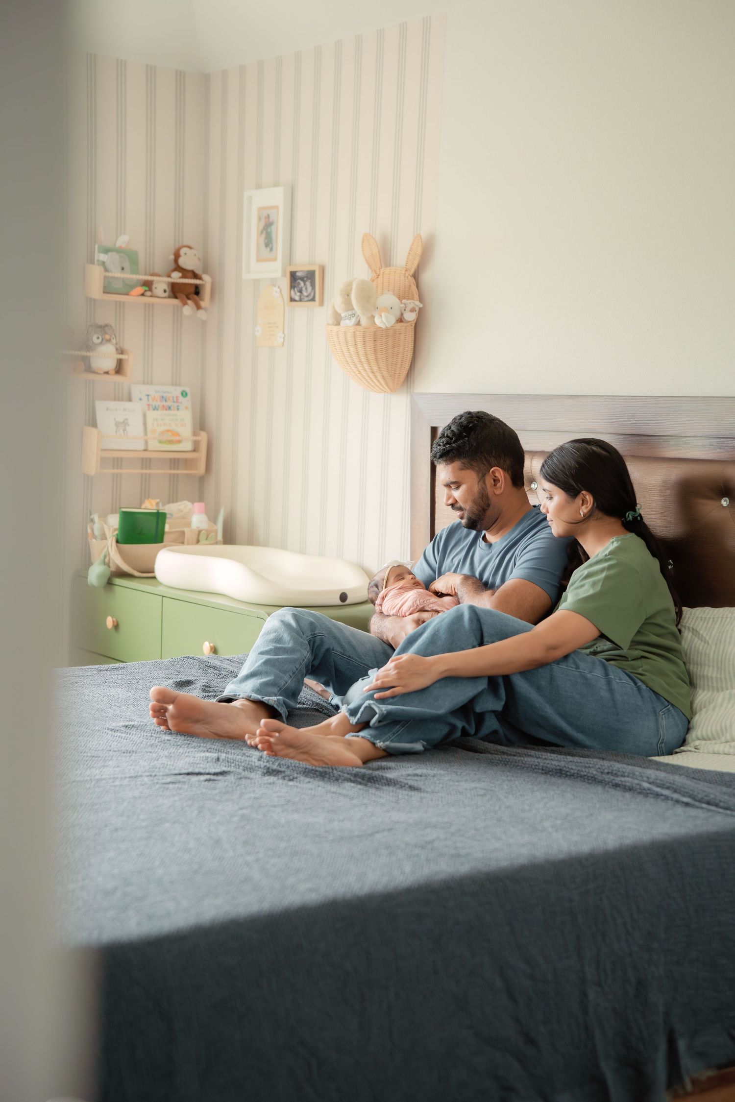 A young couple sits on a bed in a nursery, lovingly cradling their newborn baby together.