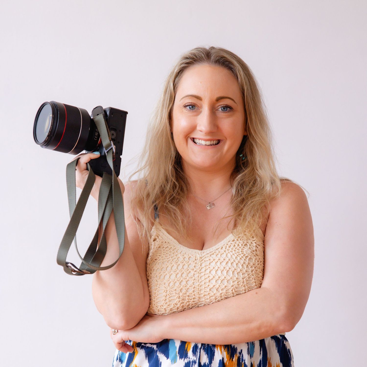 Professional photographer wearing a beige sleeveless top and blue patterned skirt holds a camera while posing for photos.