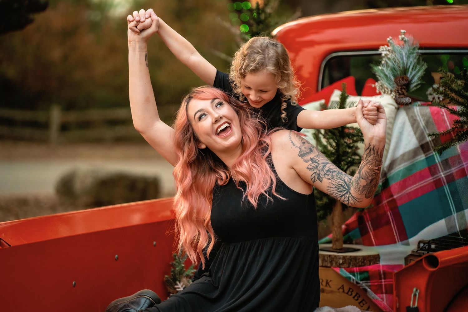 A joyful woman with pink hair and tattoos laughs with a curly-haired child in a red truck bed at Christmas.