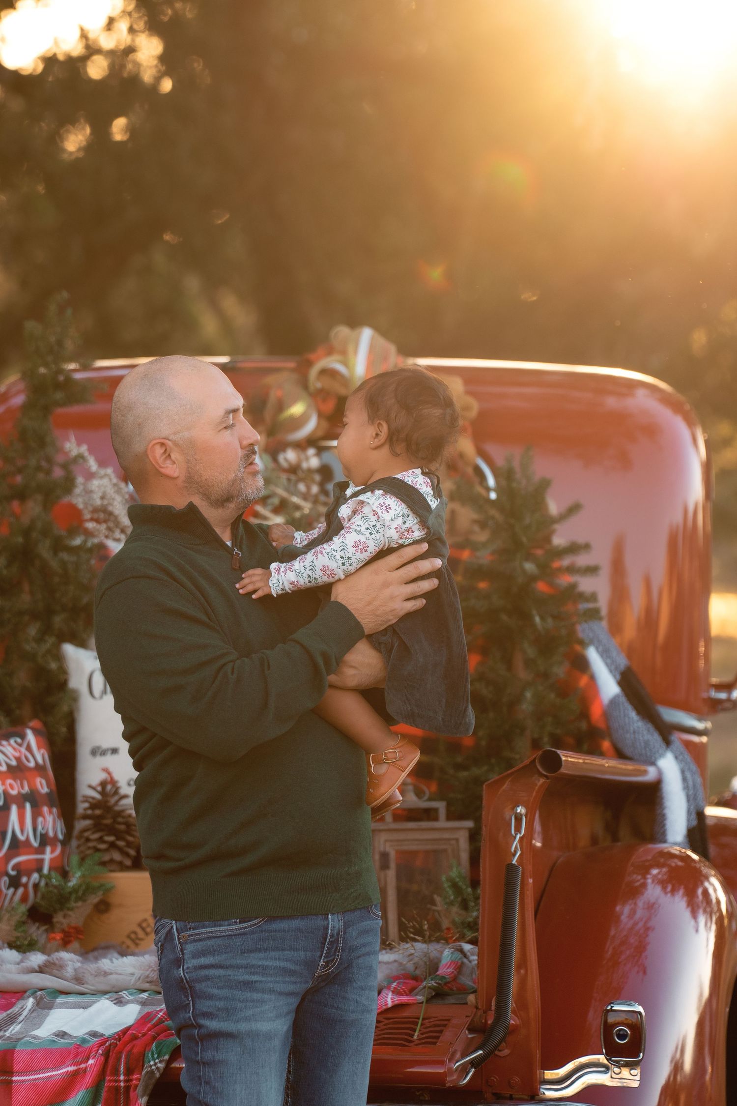 Bald man holding a baby near a red truck decorated with Christmas trees in warm golden sunset light.