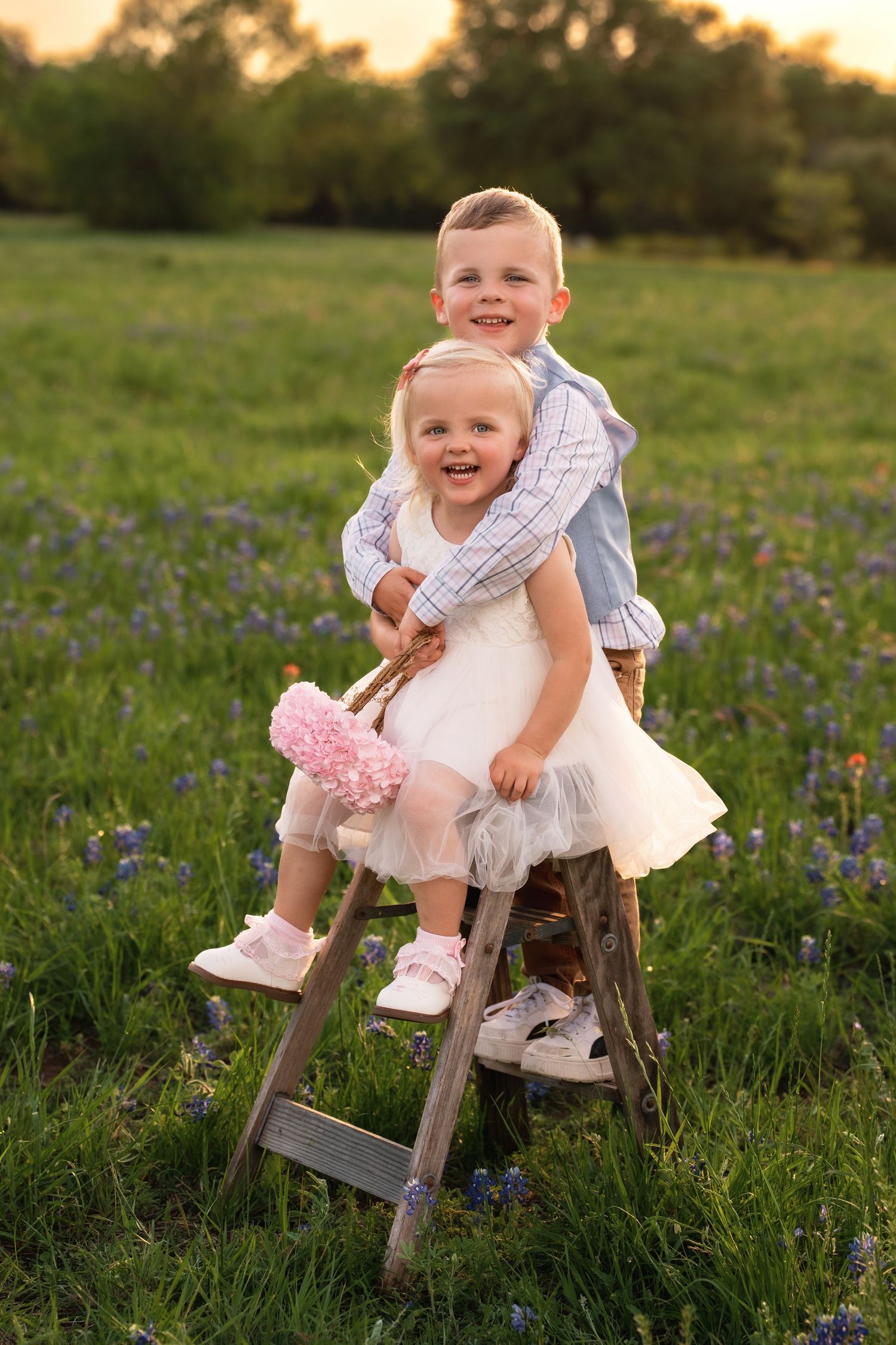 A child in a white dress and pink shoes sits on a rustic wooden ladder in a field of wildflowers at sunset.