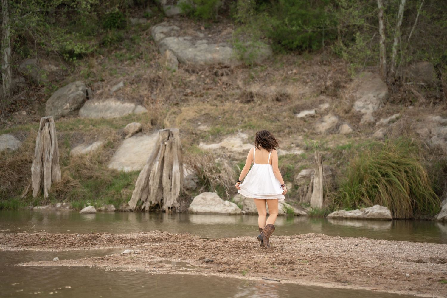 Young woman in white dress and boots walking along a rocky riverbank surrounded by trees and natural landscape.