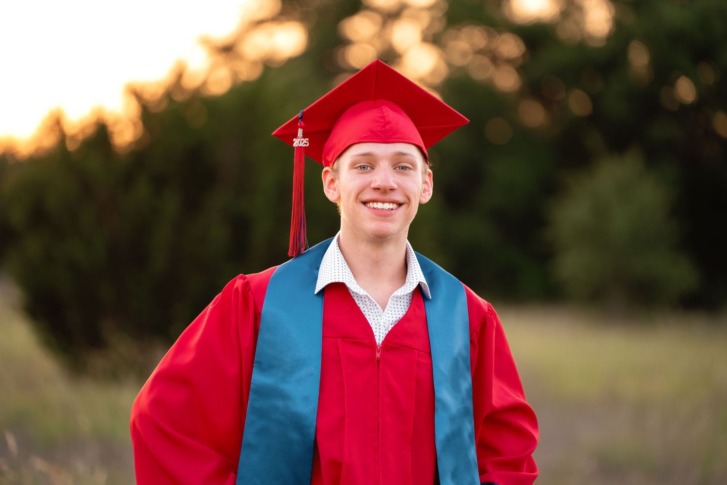 Smiling young man in red graduation cap and gown outdoors at golden hour.