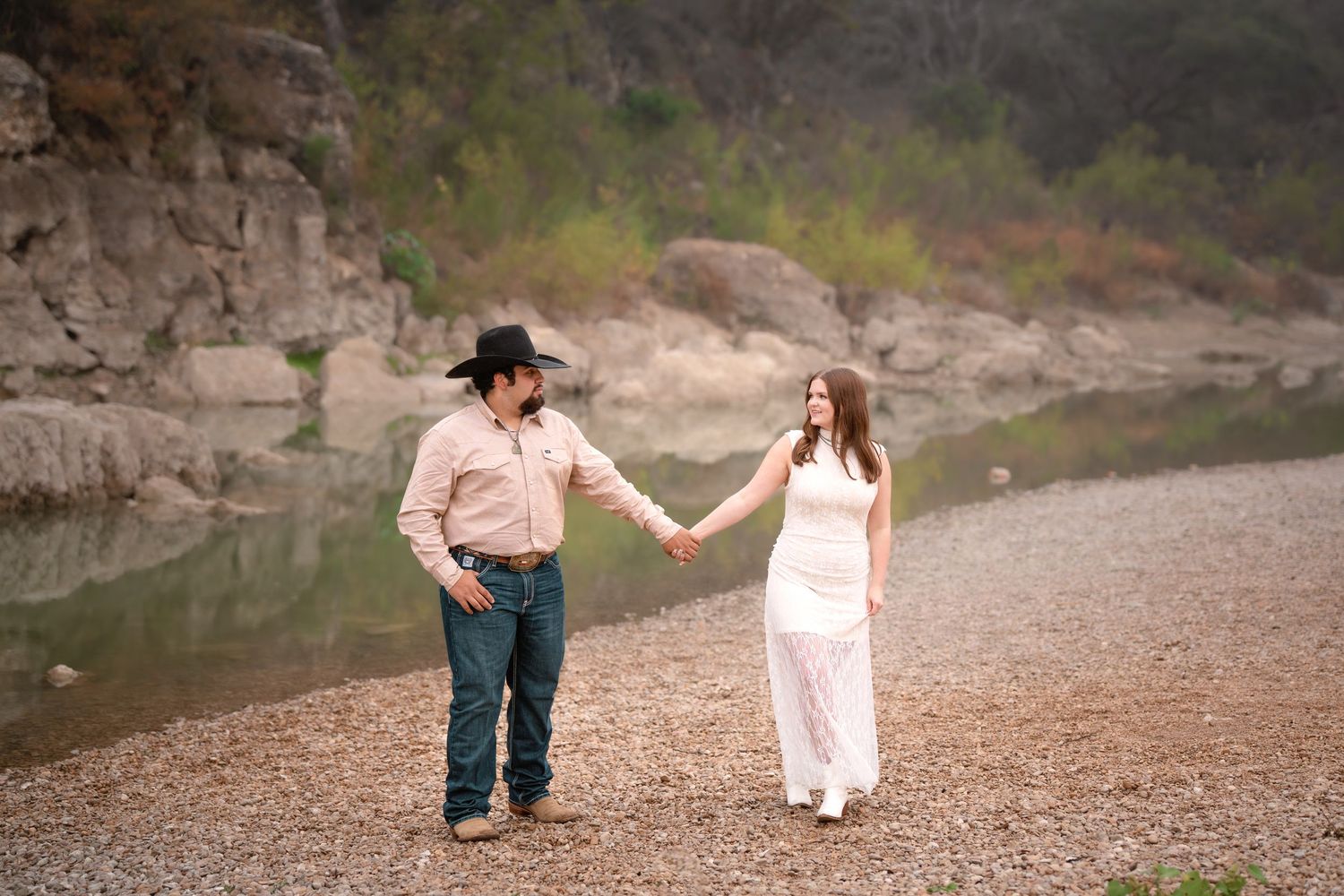 Couple holding hands on rocky riverbank, man in cowboy hat and jeans, woman in white dress, with green hills behind.