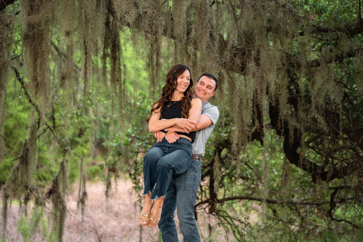 A man lifts a woman in his arms while standing among moss-draped trees in a lush green forest setting.
