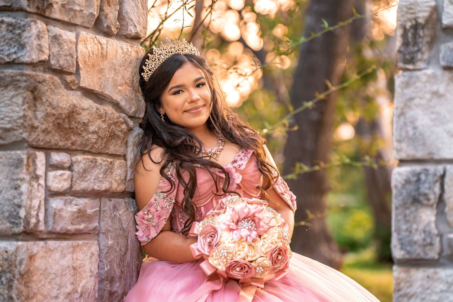 Young girl in pink quinceañera gown holding floral bouquet, wearing tiara, leaning against stone wall outdoors.