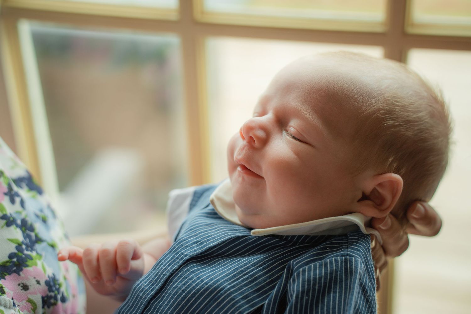 Peaceful newborn baby smiling with eyes closed, wearing a blue striped outfit, held near a bright window.