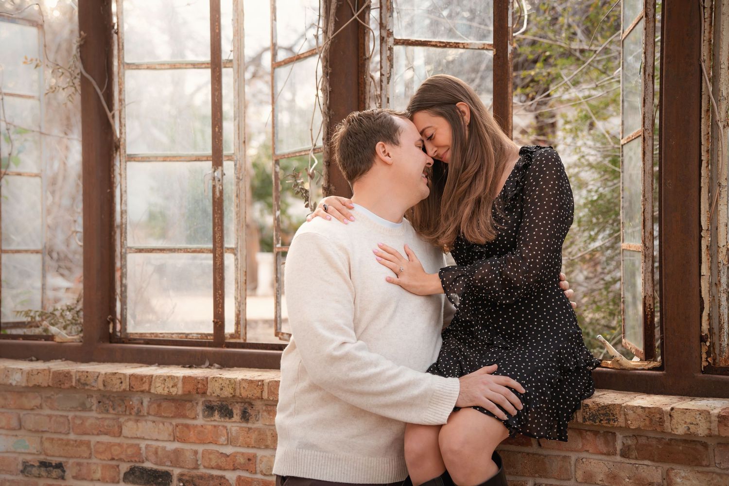 Couple sharing a romantic moment by a brick window ledge, woman in floral dress seated while man in white sweater leans in.