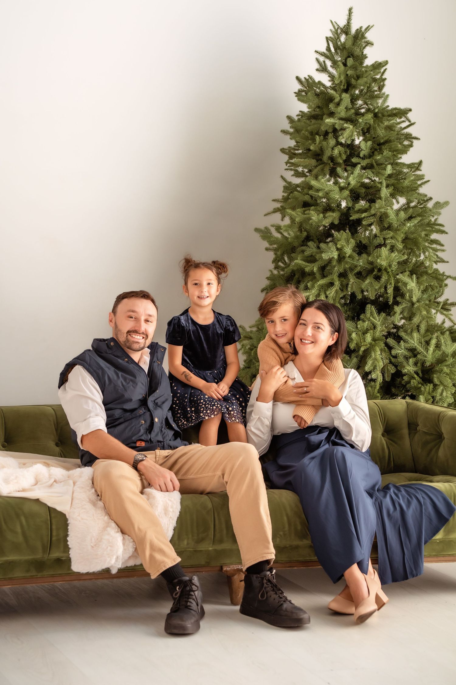 Happy family of four posing on green velvet sofa in front of Christmas tree in festive holiday photo.