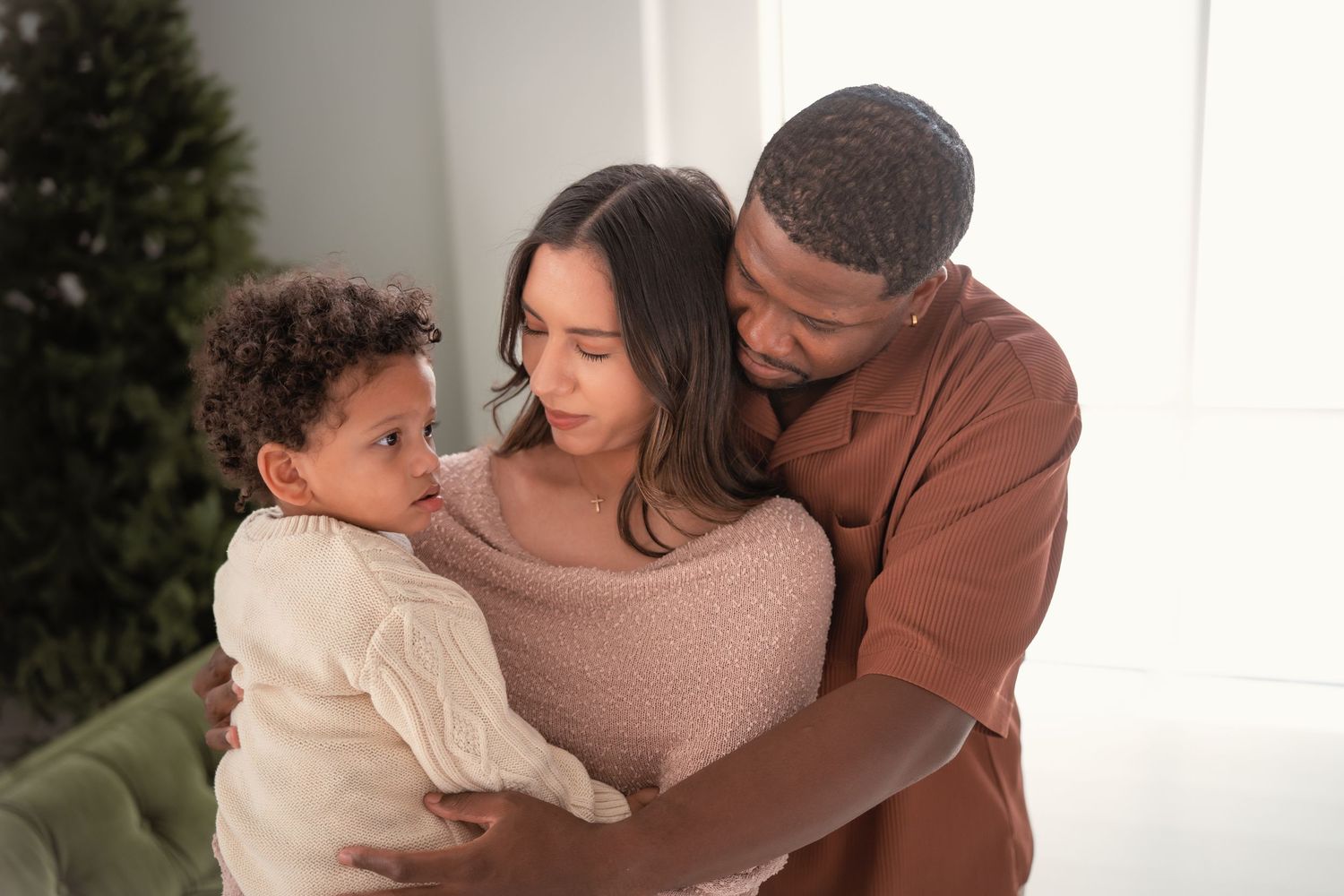 A loving multiracial family with parents tenderly embracing their young curly-haired toddler indoors.