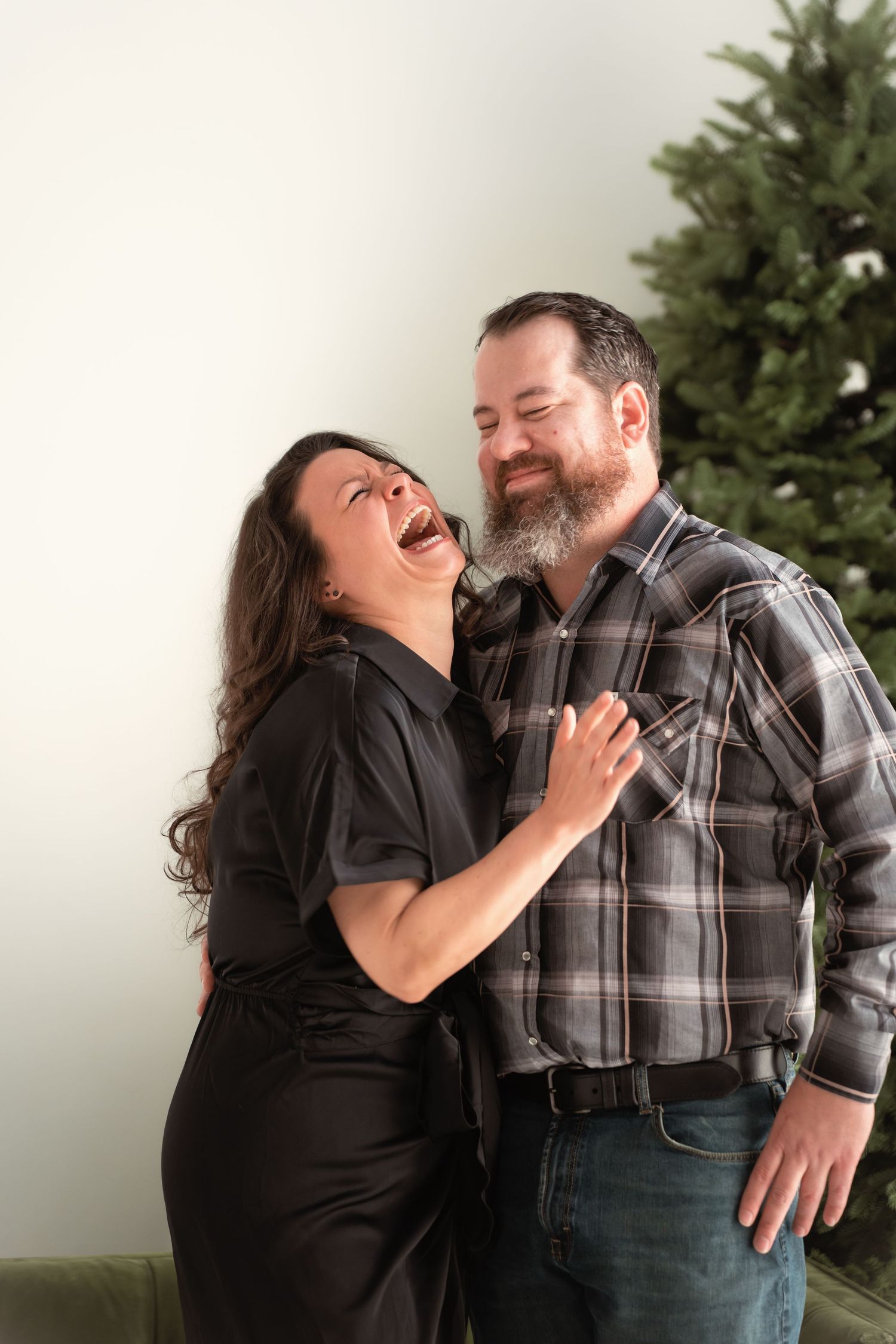 Couple laughing and embracing in front of a Christmas tree, woman in black jumpsuit and man in plaid shirt.