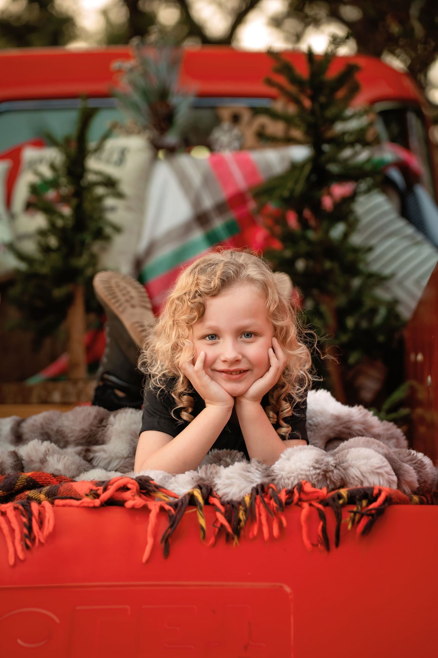 Smiling curly-haired child leaning on red truck tailgate with Christmas trees and plaid blanket in festive holiday setting.