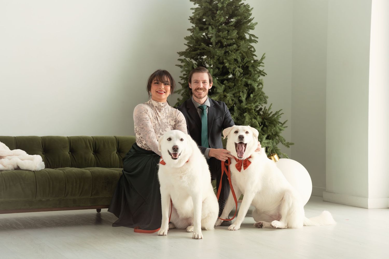 Couple posing with two white dogs near a decorated Christmas tree on a green velvet sofa.