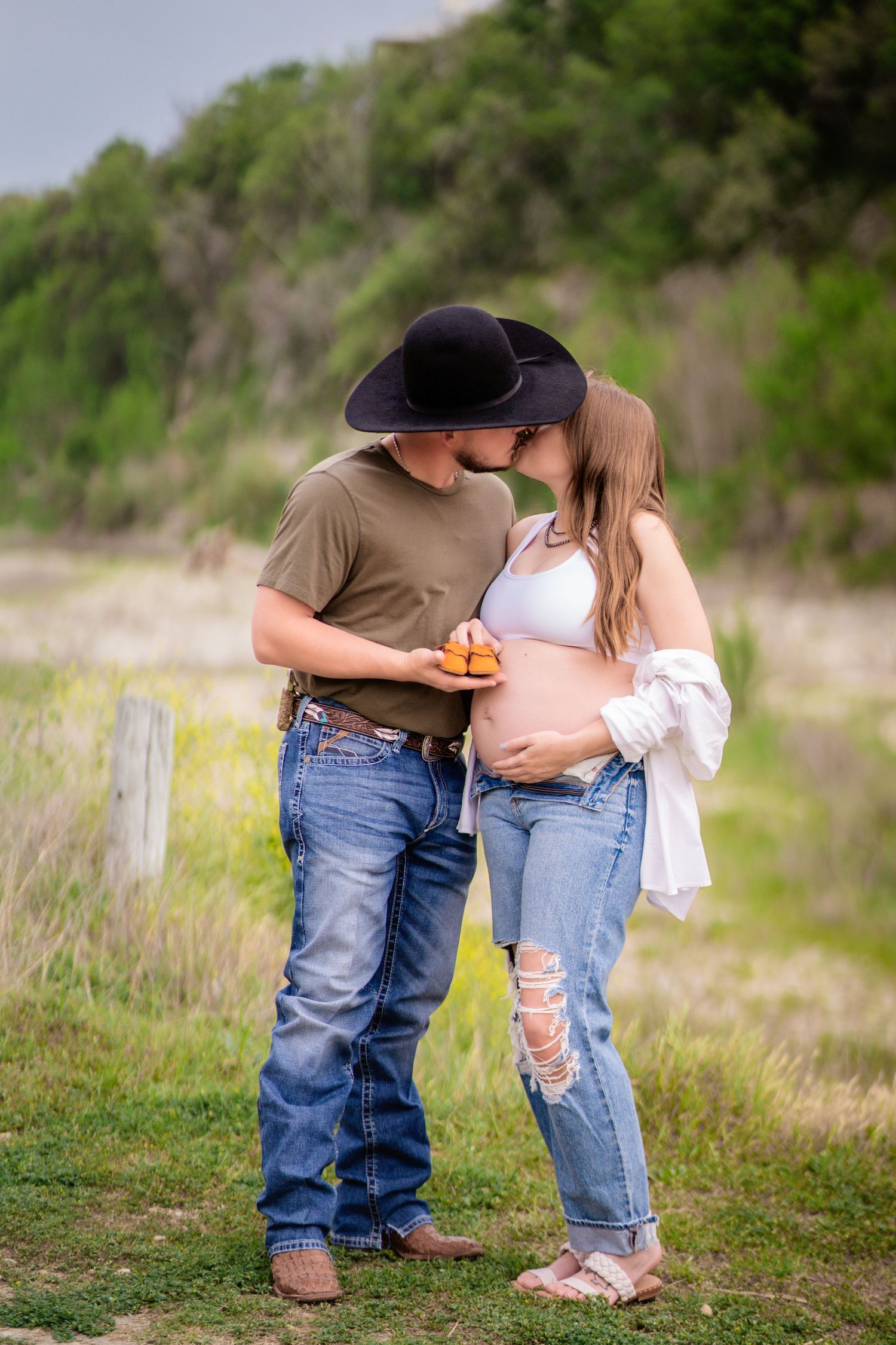 Couple sharing a kiss outdoors, man in cowboy hat holding peaches beside pregnant woman in ripped jeans and white top.