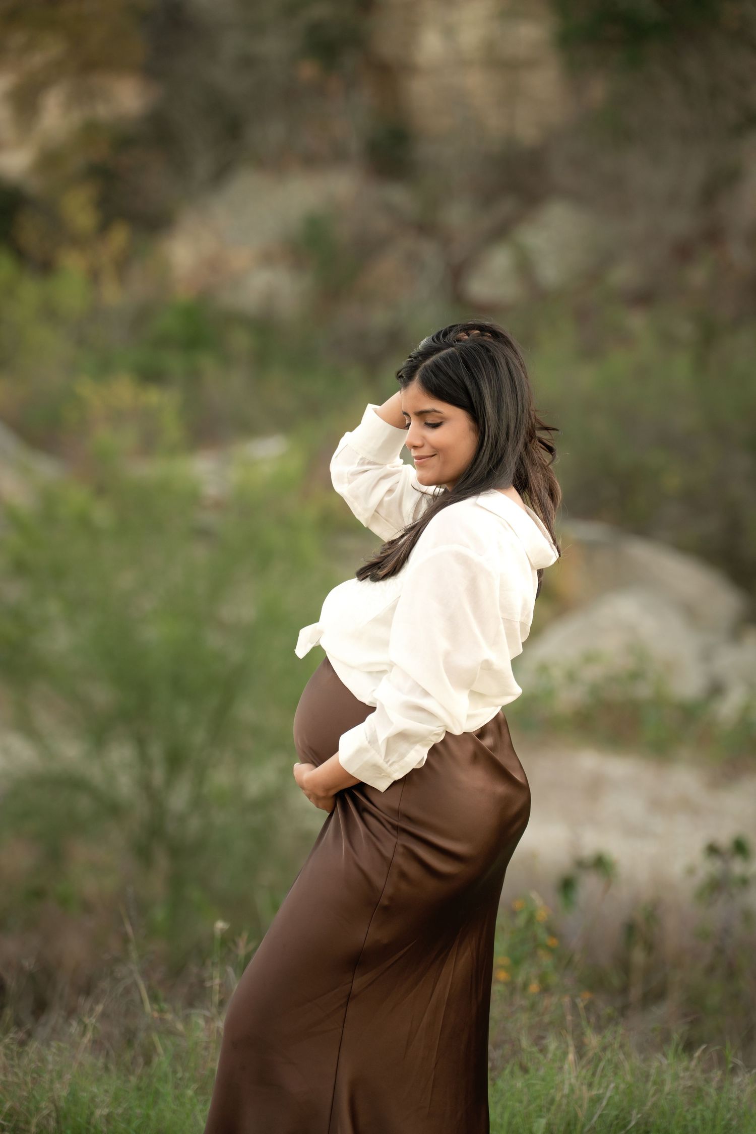 Pregnant woman in white blouse and brown skirt poses outdoors in nature, cradling her baby bump with a serene smile.