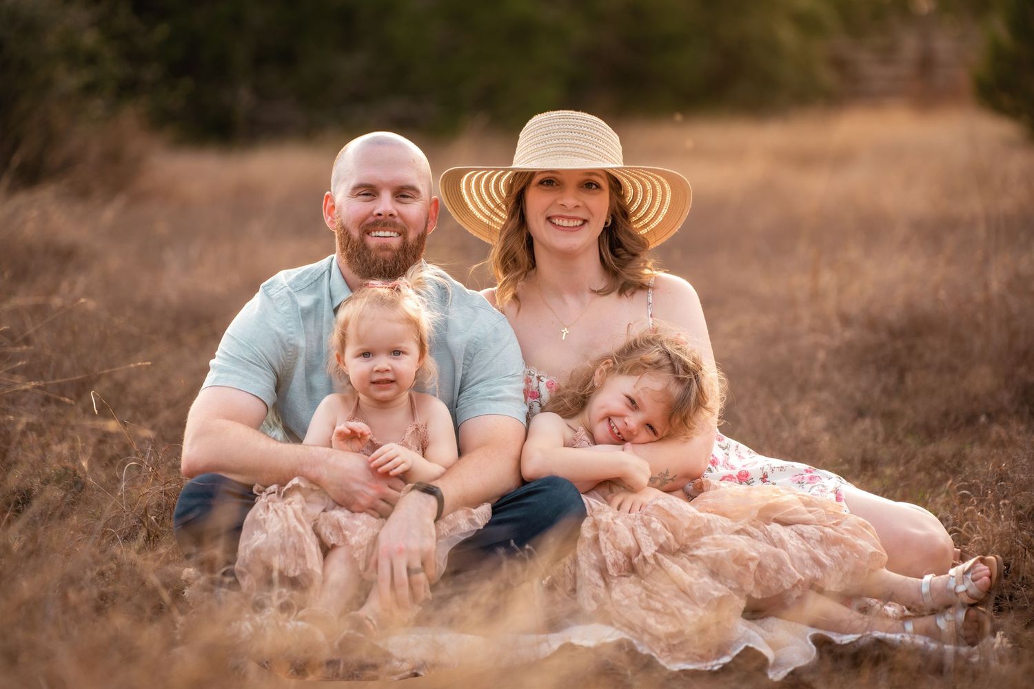 Family of four smiling outdoors in golden field, parents with two young daughters in floral dresses, warm sunny day.