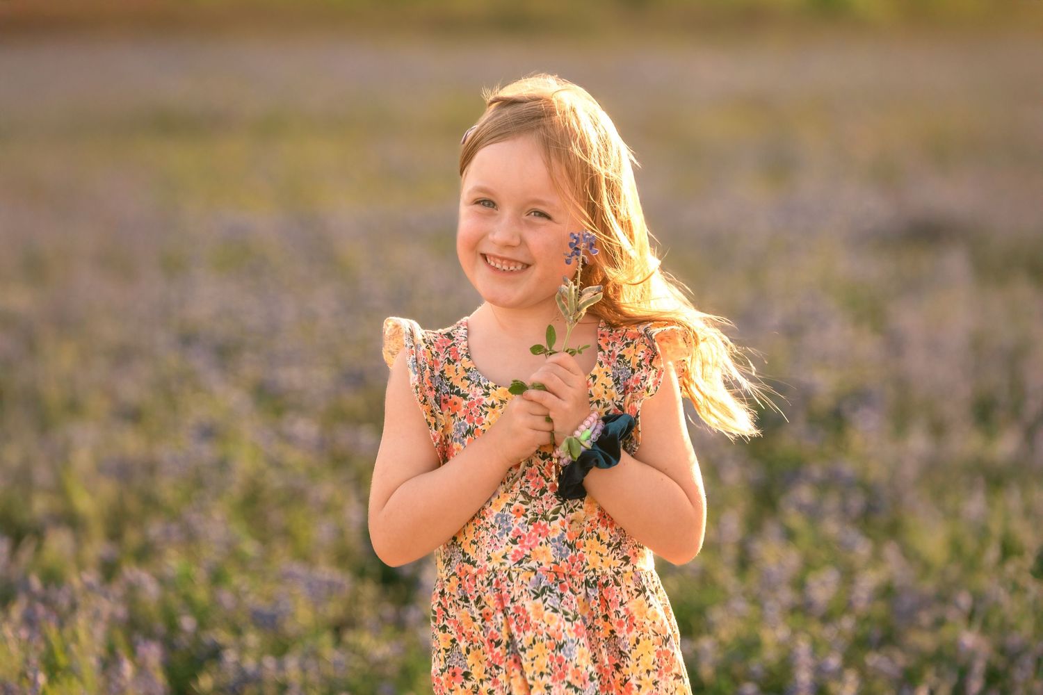 A young person in a floral dress stands in a blooming lavender field at golden hour with windswept hair.