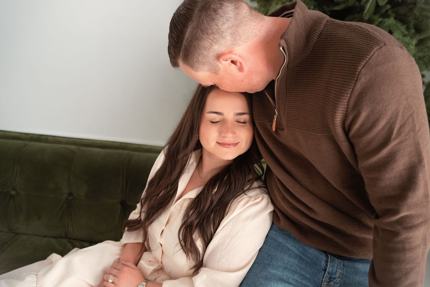 Couple shares tender moment on green velvet sofa, man kissing woman's forehead affectionately.