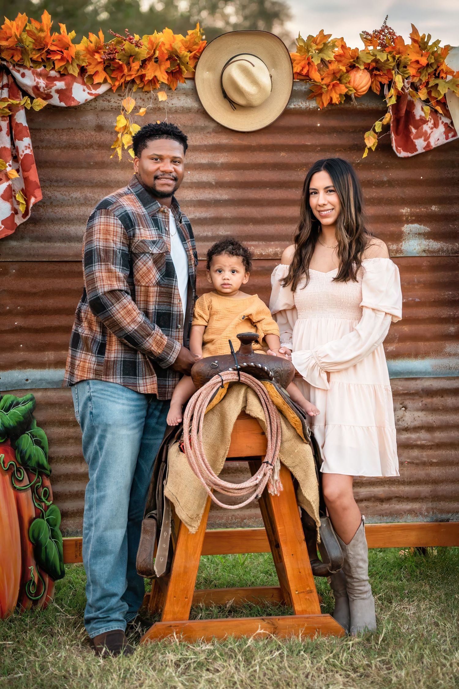 Family poses together in warm autumn setting with wooden horse and rustic corrugated metal wall backdrop decorated with fall leaves.