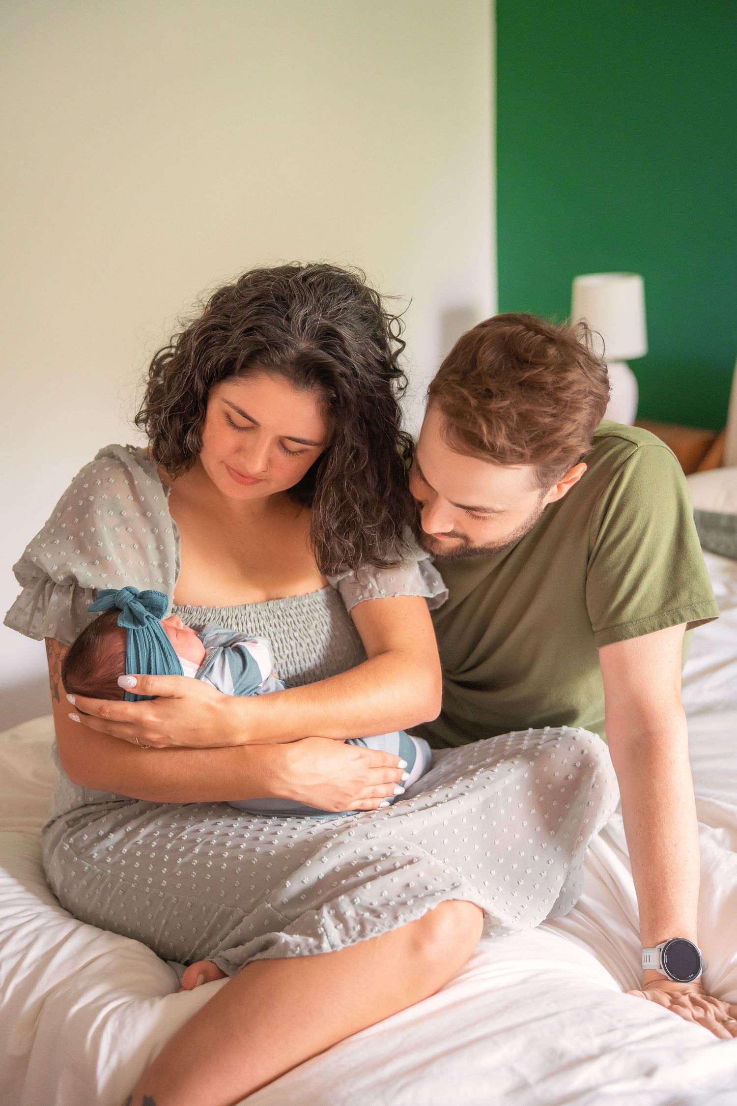 New parents sitting on bed, mother cradling newborn baby wrapped in blue blanket, father leaning in to admire.