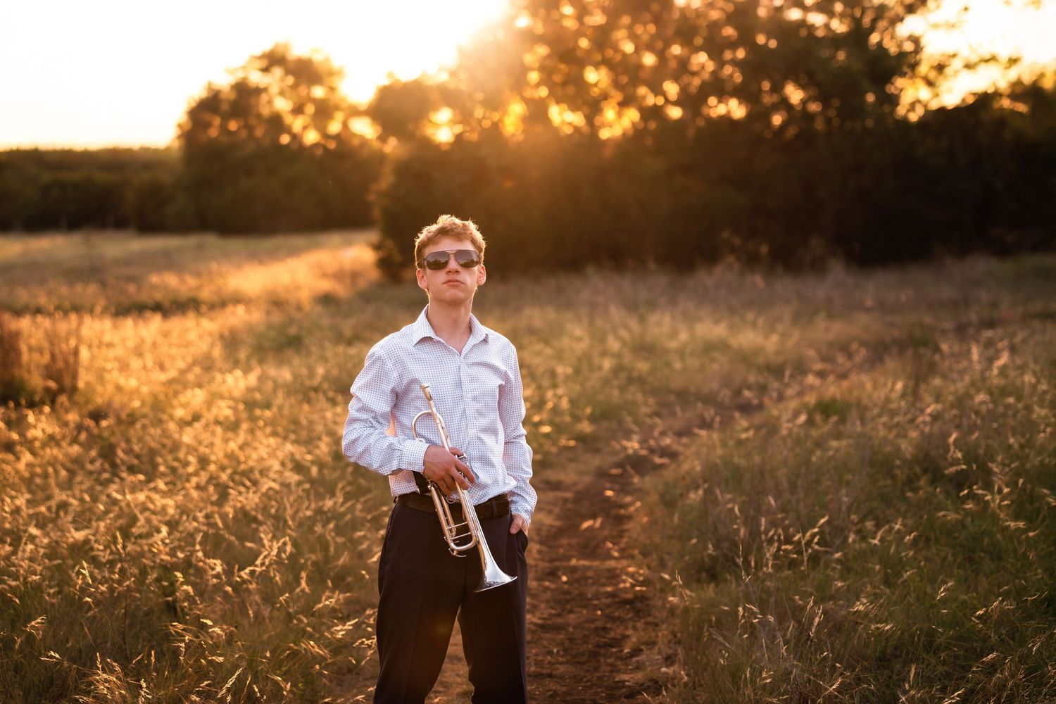 Young boy in white shirt holding a trumpet in a golden sunlit field at sunset wearing sunglasses.