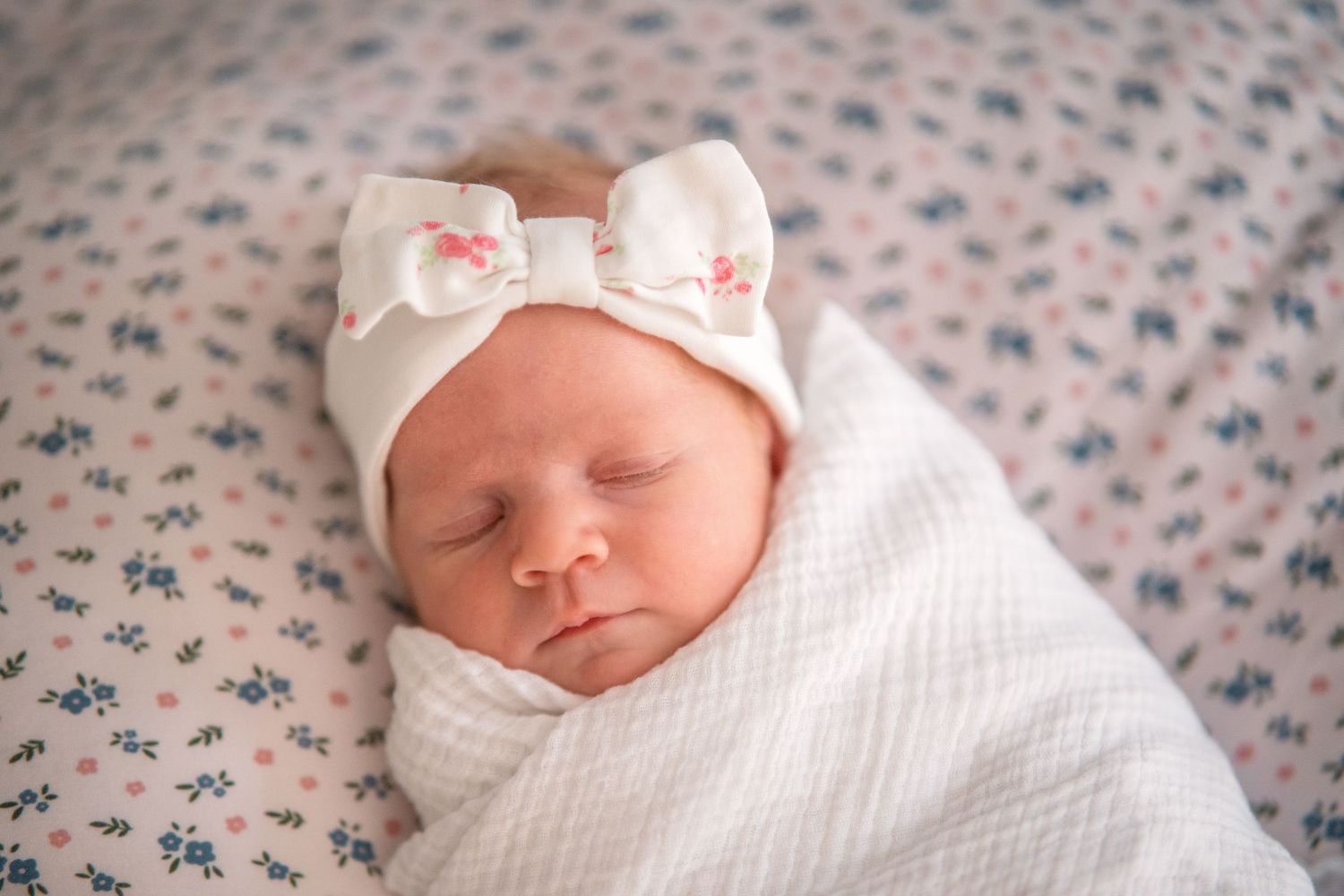 Peaceful newborn baby girl swaddled in white, wearing a floral bow headband, sleeping on a floral blanket.