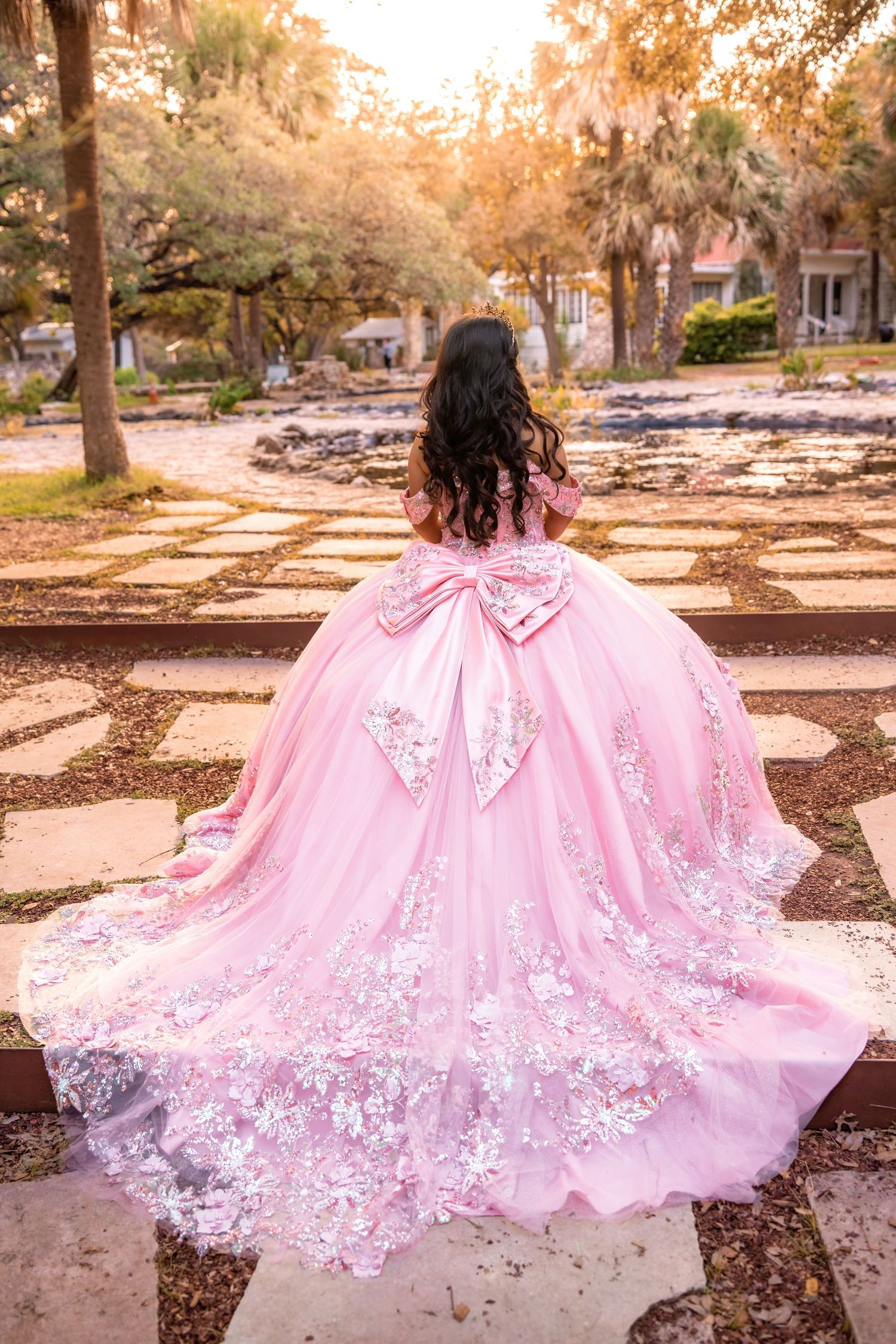 Young woman in a pink ballgown with lace floral details and large bow, standing in a garden pathway.