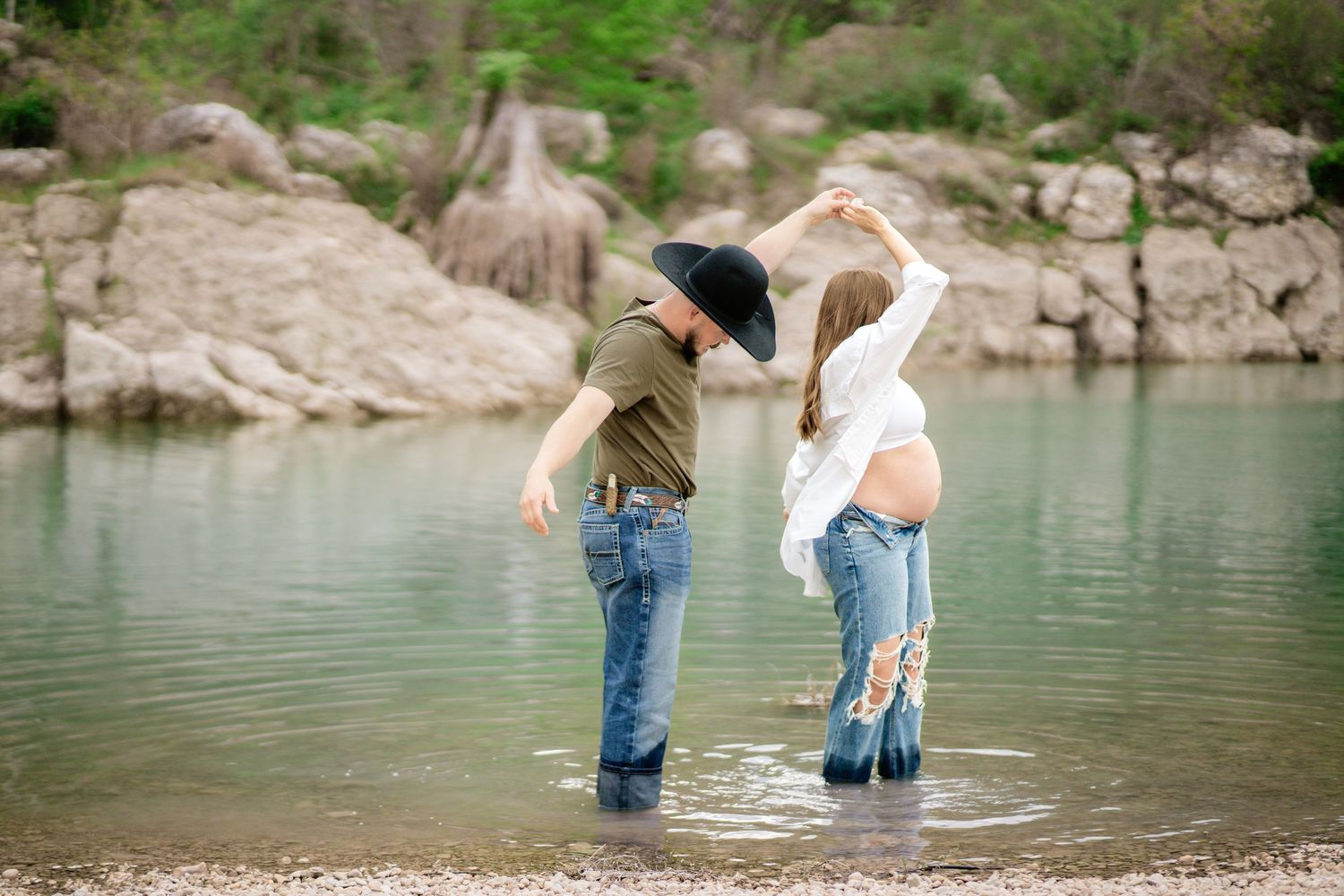 Couple dancing in shallow water, pregnant woman in white top and jeans, man in cowboy hat, rocky shore backdrop.