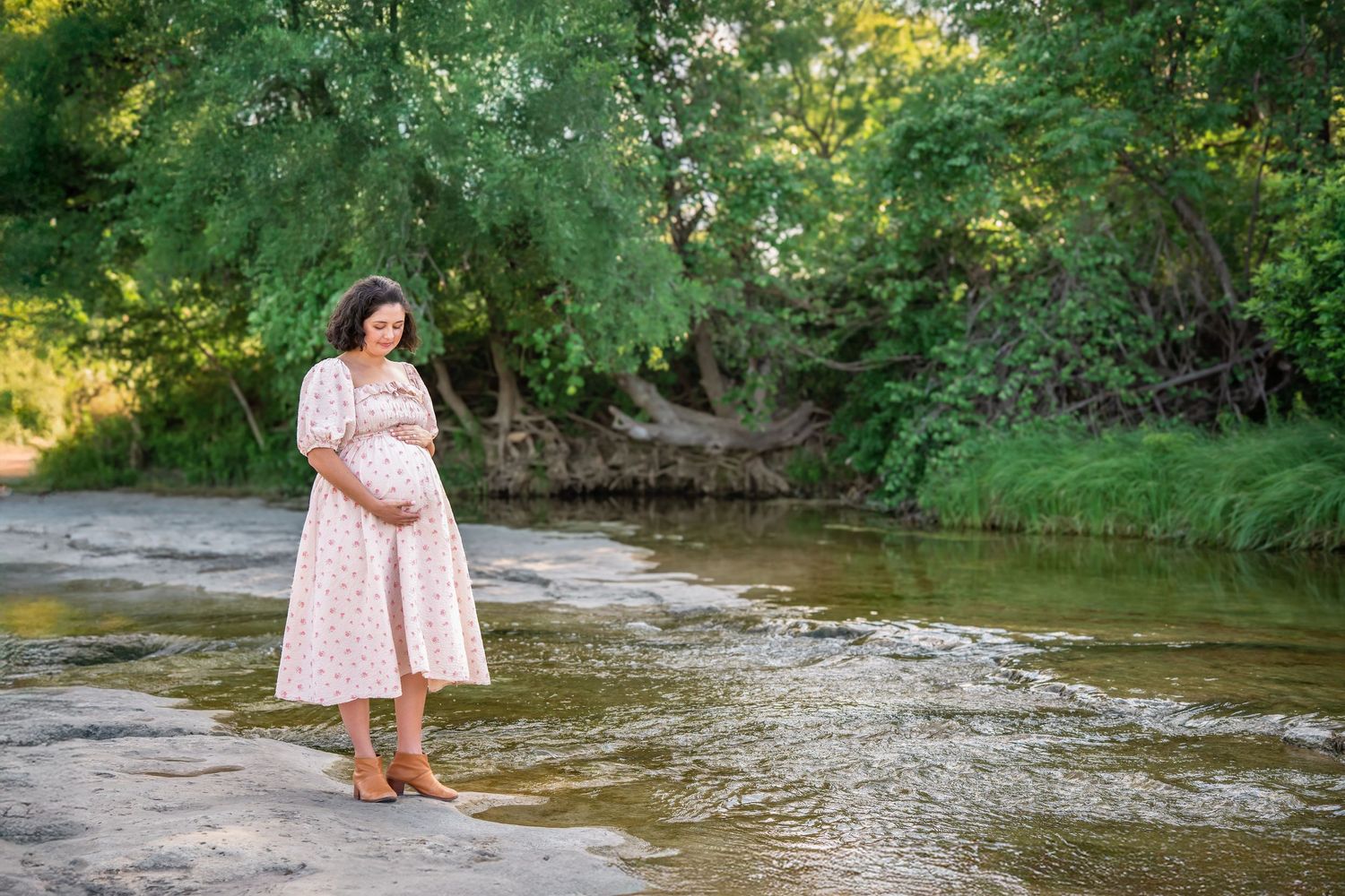 Pregnant woman in floral dress standing barefoot by a peaceful creek surrounded by lush green trees.