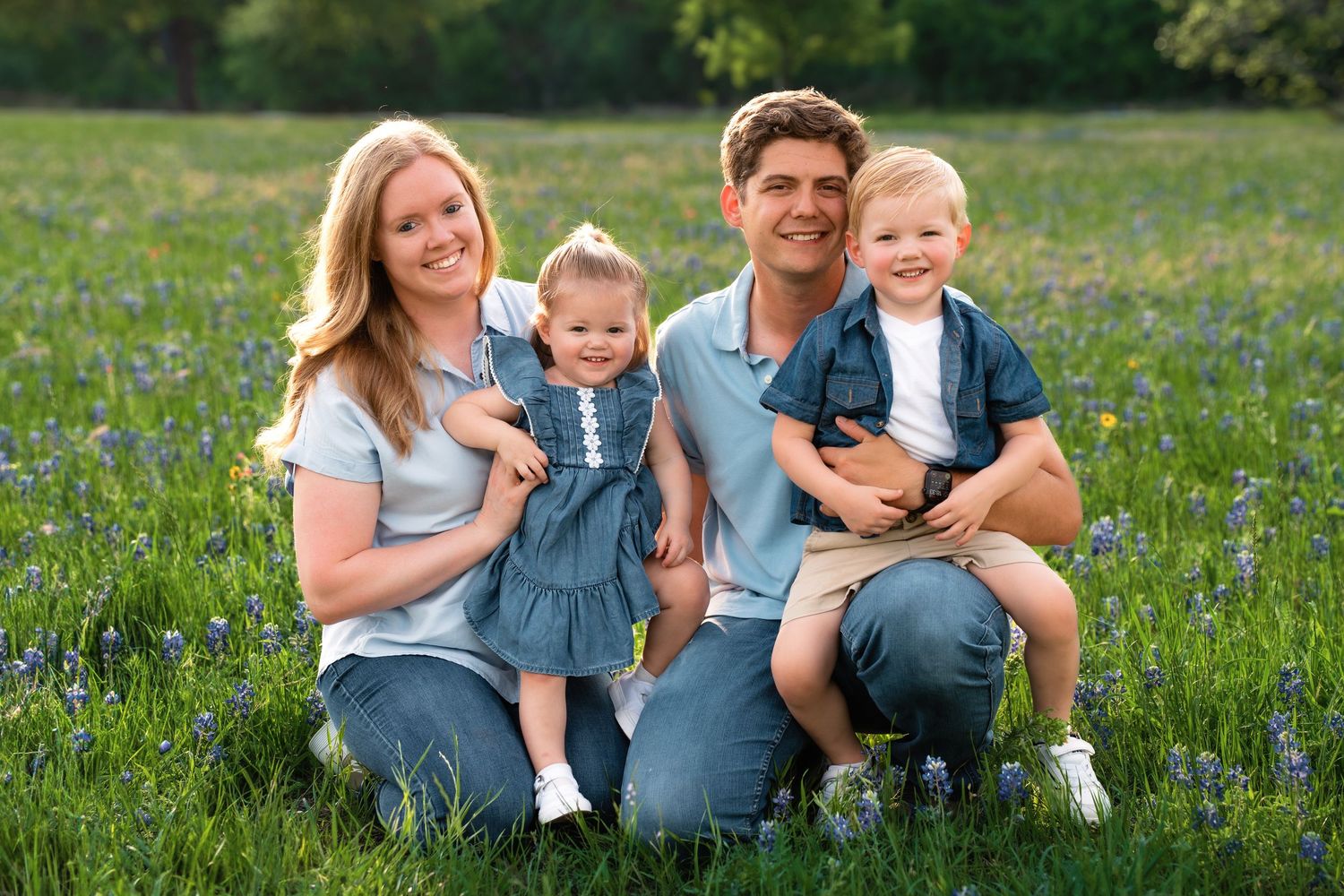 A family sits together in a blooming wildflower meadow wearing coordinated blue and white outfits on a sunny day.