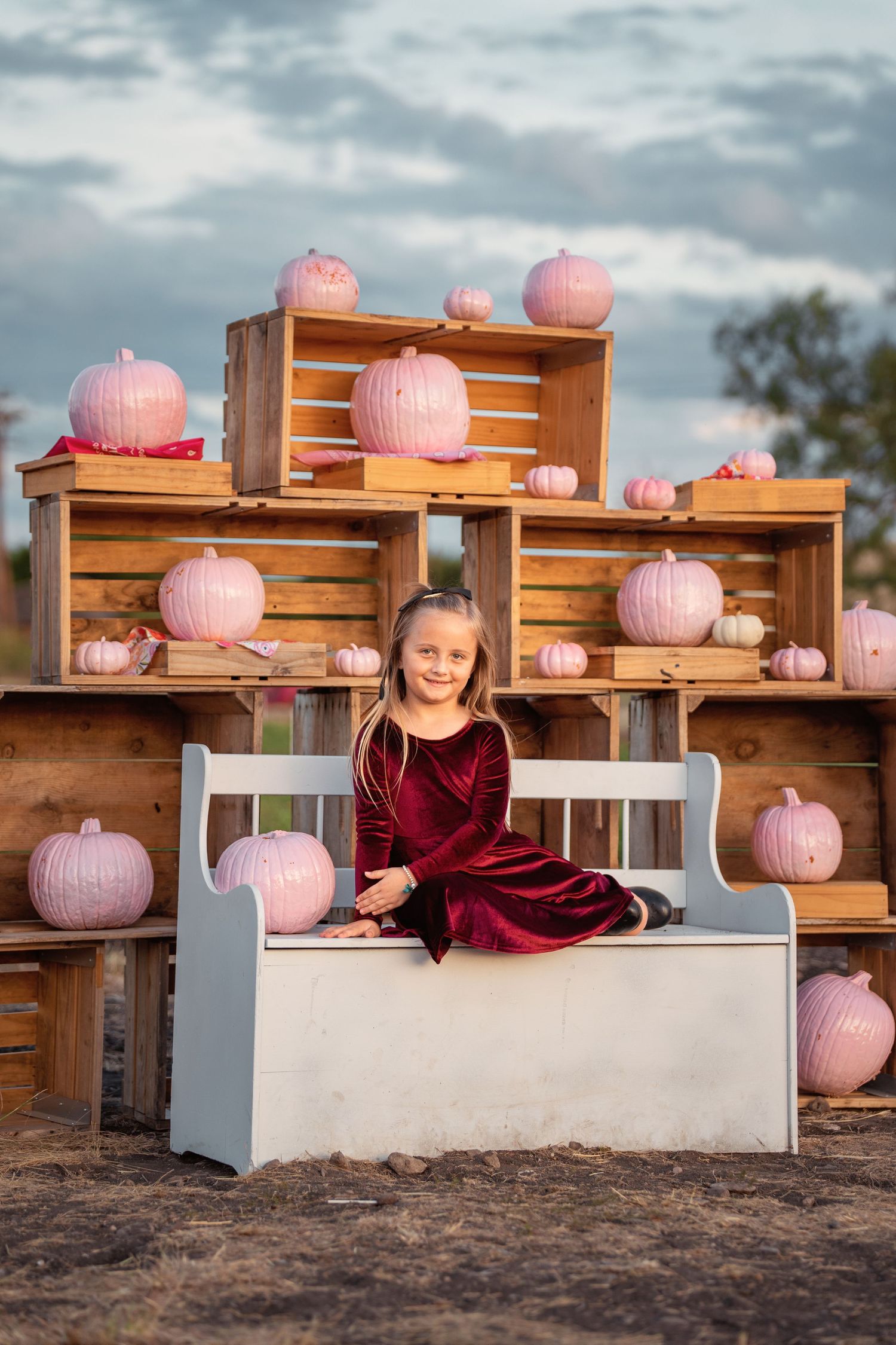 A young girl in a burgundy velvet dress sits on a white bench surrounded by pink pumpkins and wooden crates outdoors.