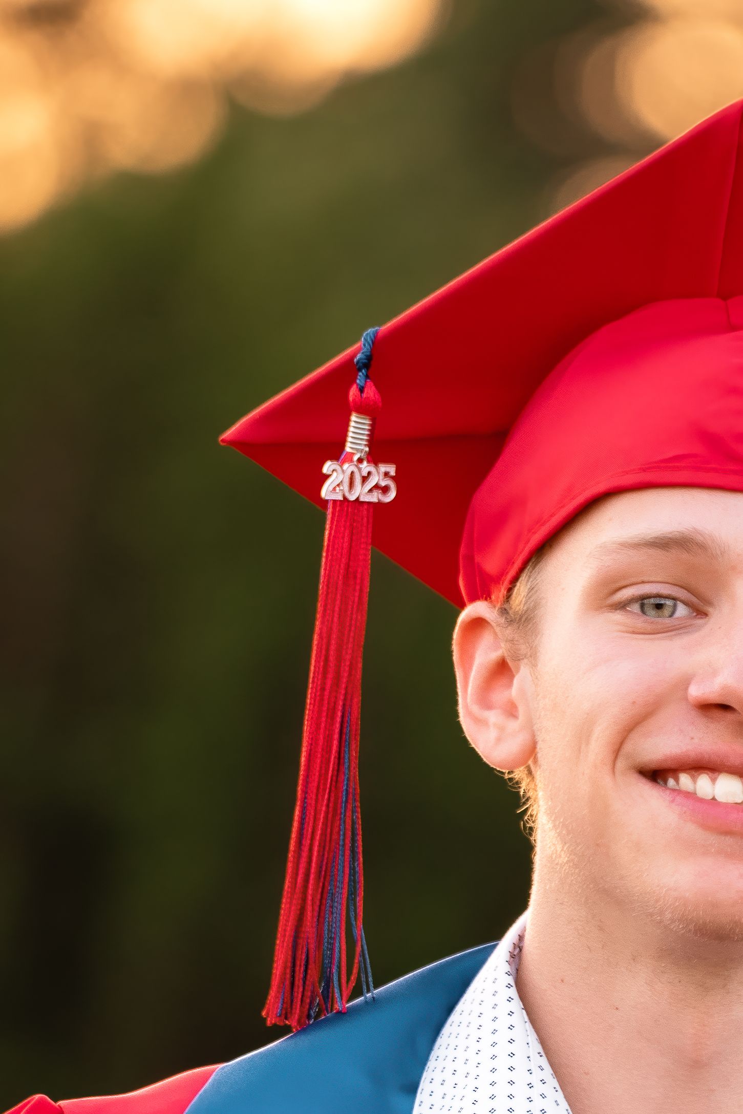 Smiling young man wearing a red 2025 graduation cap with tassel outdoors during golden hour.