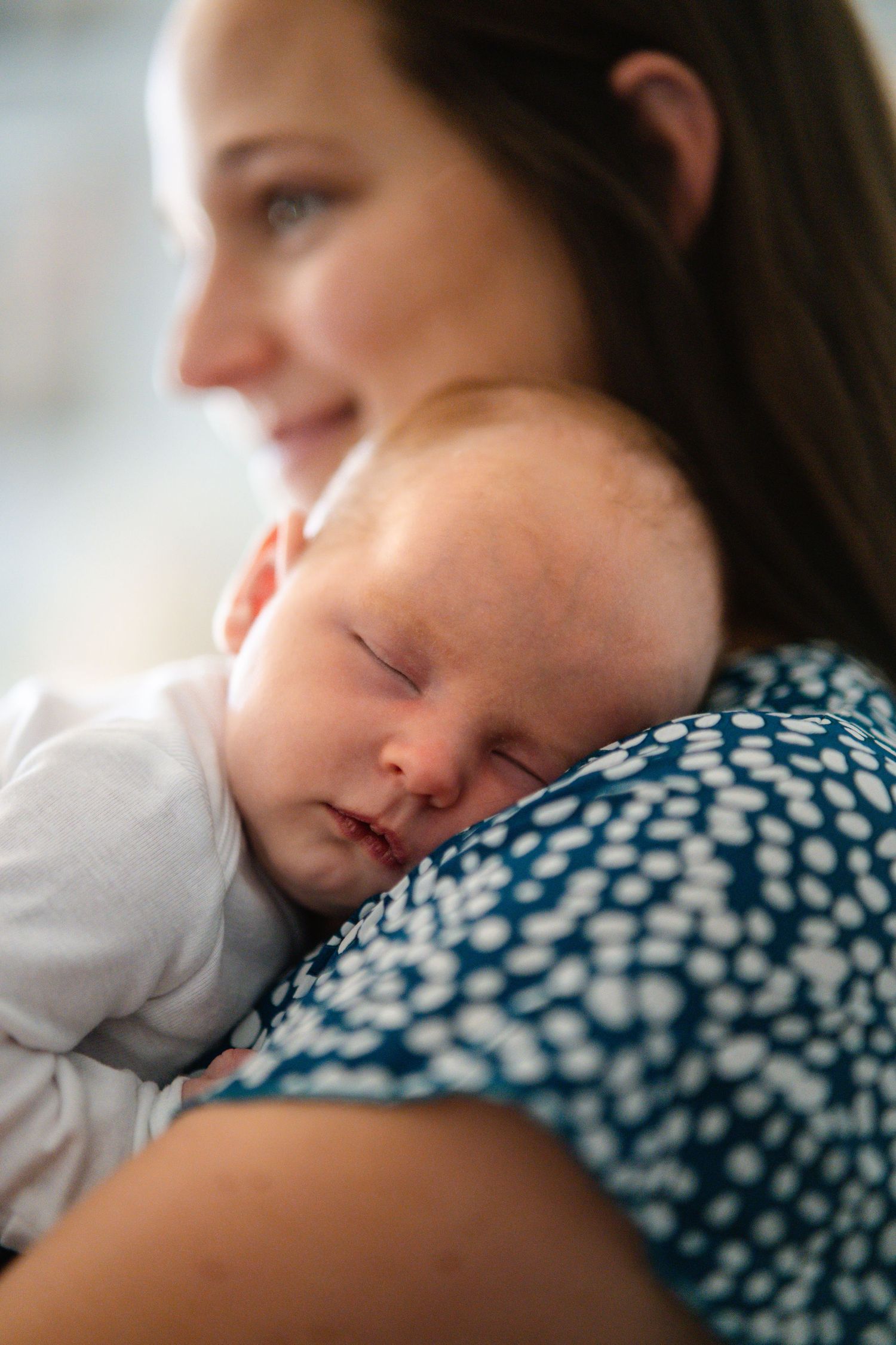 A newborn baby sleeping peacefully on a smiling mother's shoulder, dressed in a blue patterned top.