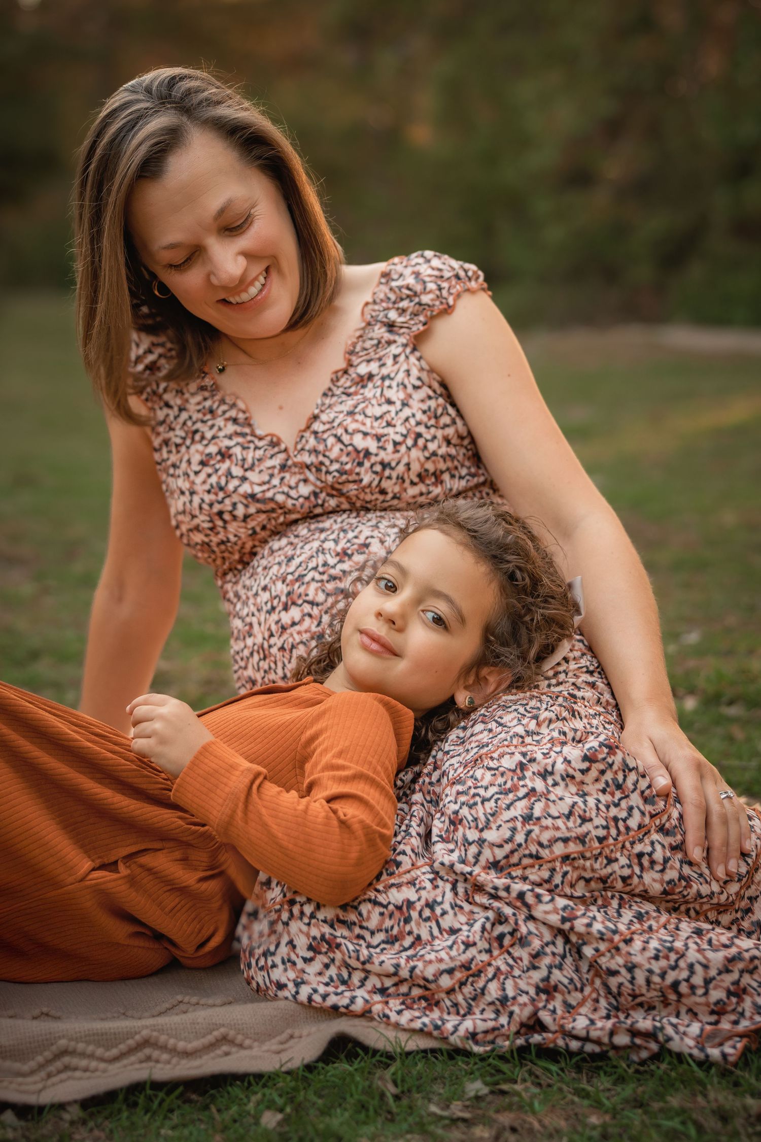 Pregnant woman in floral dress smiling as young girl rests head on her baby bump outdoors in autumn park.